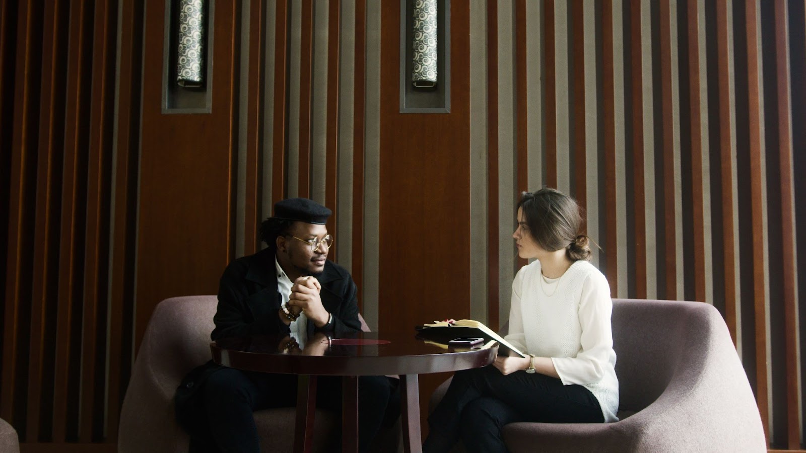 An image of a man and a woman sitting on opposite facing chairs, seemingly engaged in conversation. The woman is wearing a white blouse and holding a notebook. The man is wearing black and listening to the woman.