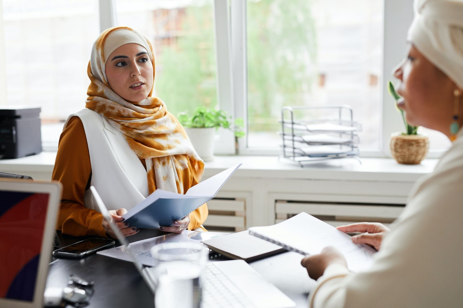 A picture of two women sitting across from each other in an office, holding papers and having a conversation.