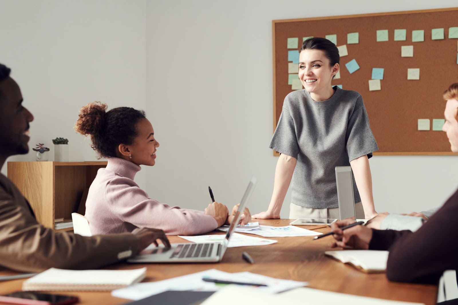 Photo of a group of professionals having an informal meeting around a table with a corkboard in the background.