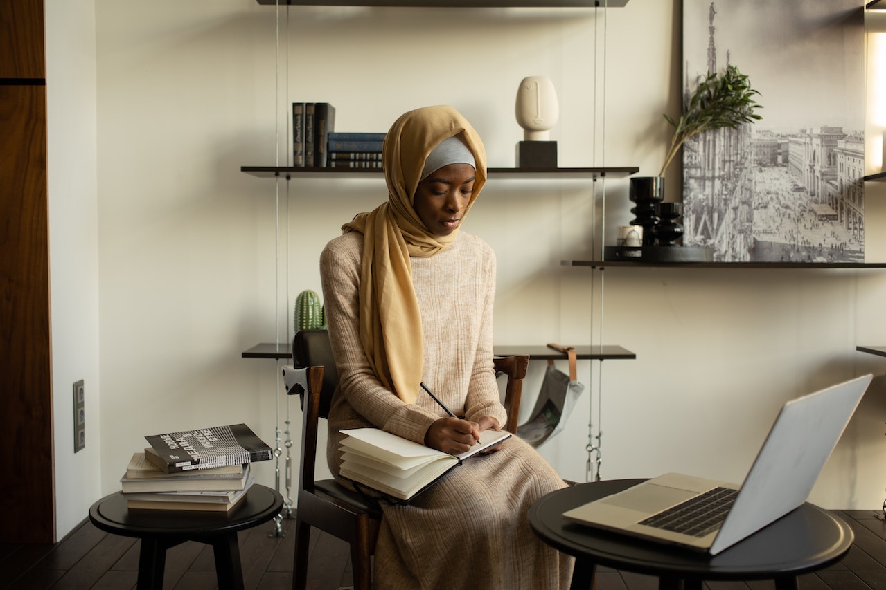 A photograph of an individual sitting on a chair, taking notes, with a laptop in front of them. 